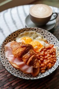 A hearty plate of English breakfast with eggs, beans, ham croissant, and coffee on a patterned plate.